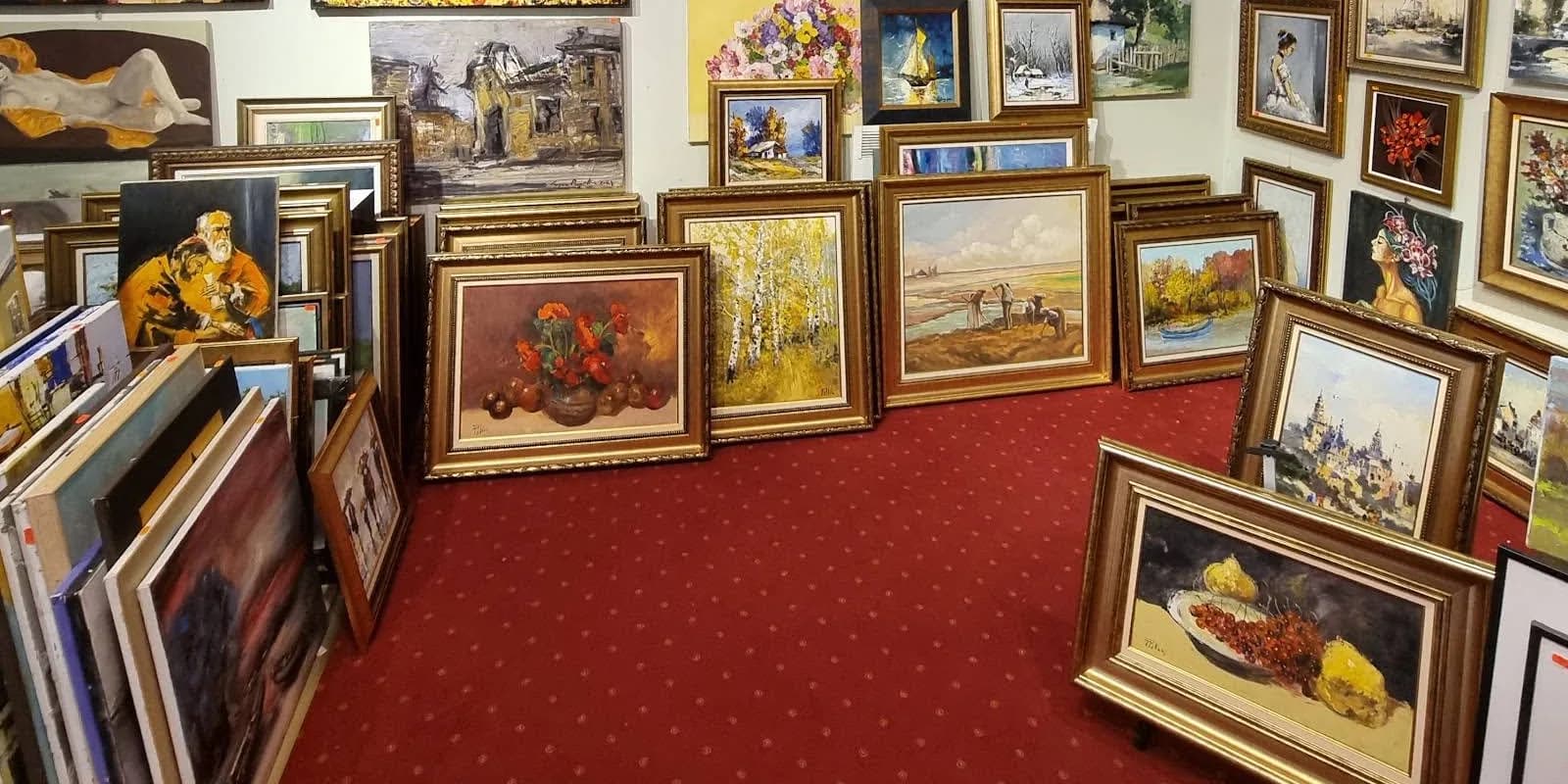 Lucrări înrămate așezate pe muchie pe podeaua galeriei, sprijinite una de alta în teancuri, pe un covor roșu