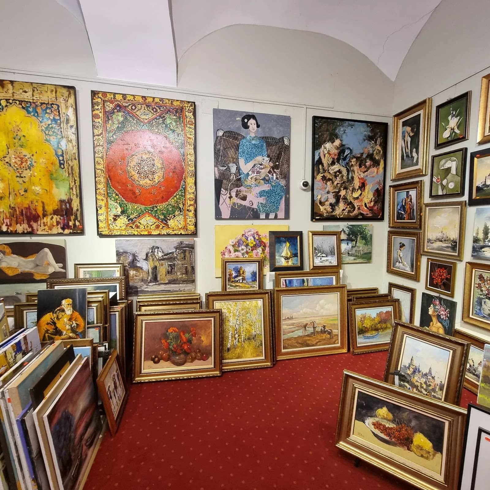 Sala galeriei sub bolți albe: tablouri agățate pe pereți și altele sprijinite unele de altele pe covorul roșu