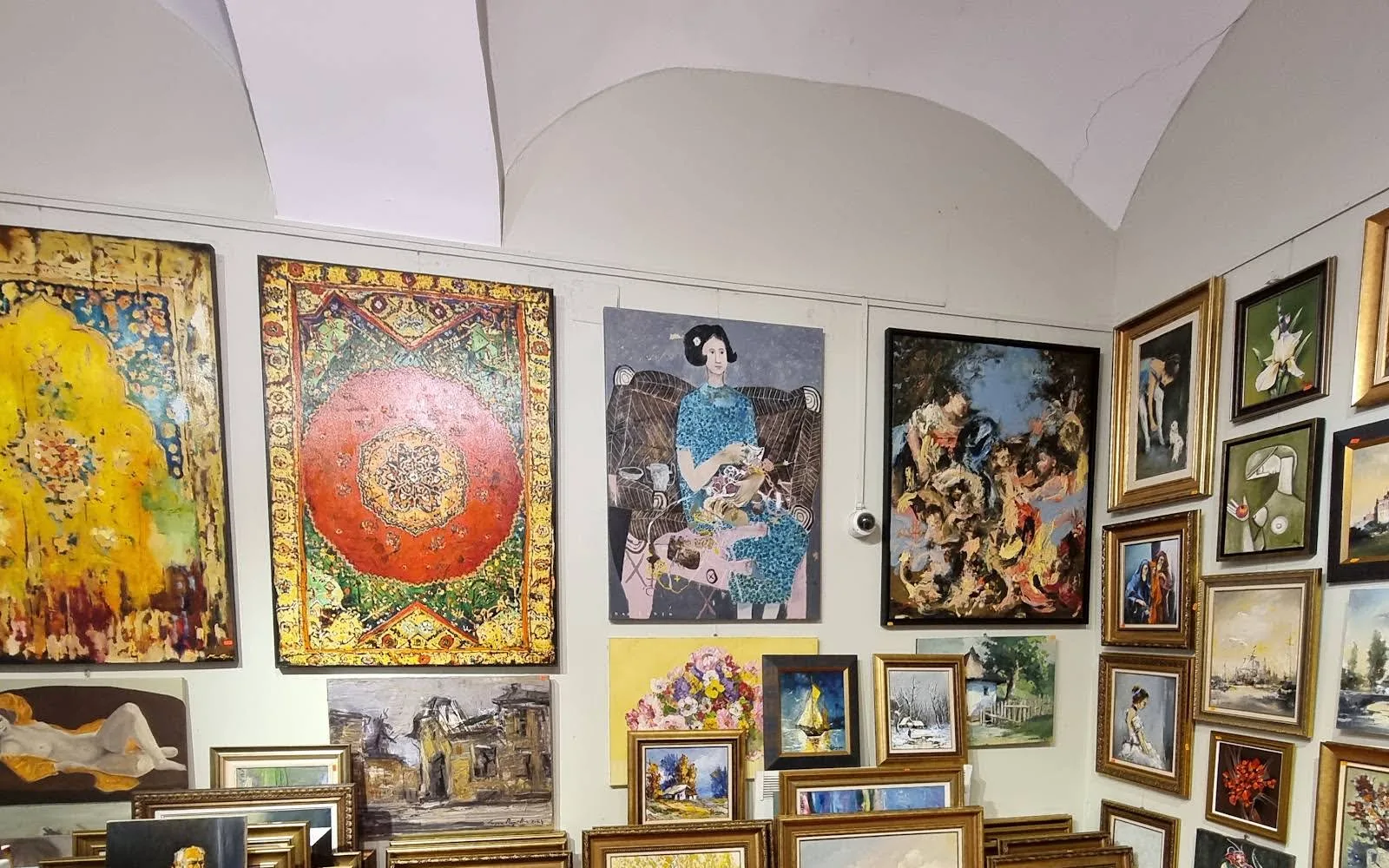 Încăpere cu tavan boltit alb și pereți plini de tablouri, de la covoare pictate în galben și roșu până la peisaje mici în rame aurii